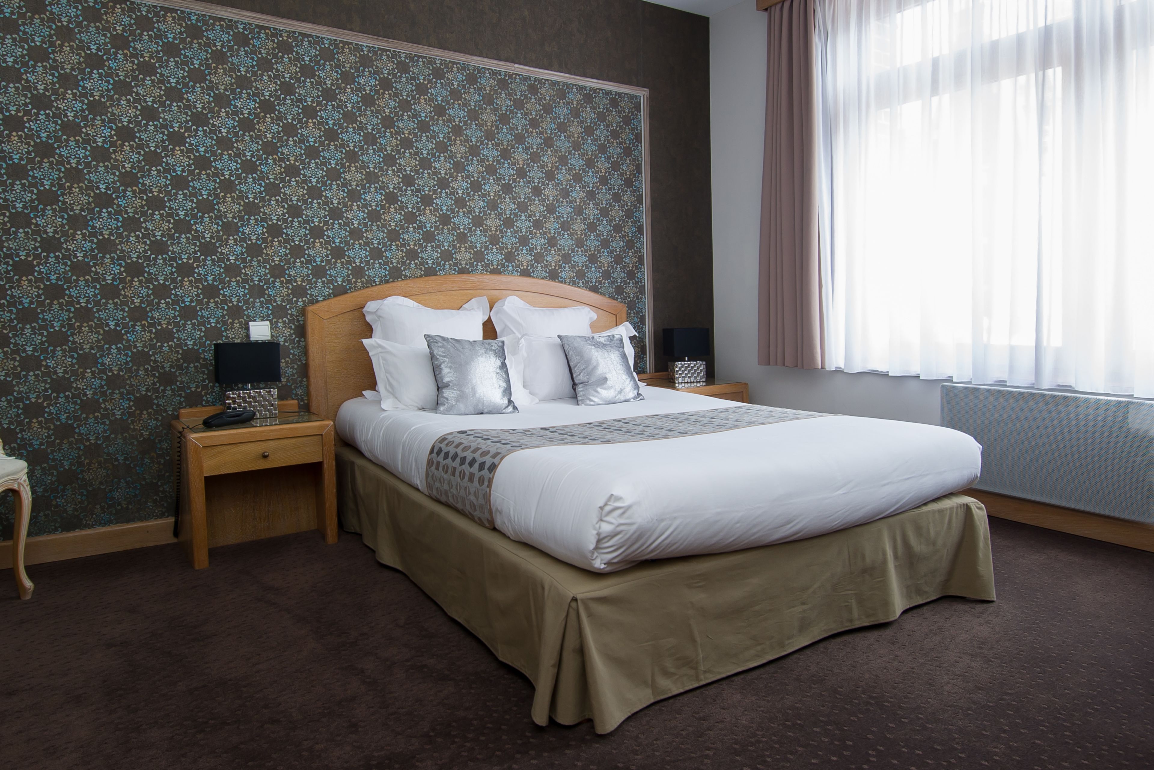 A neatly made bed with a wooden headboard, a beige bed skirt, and multiple pillows. There are two bedside tables with lamps, a small nightstand, and a patterned wallpaper on the wall.