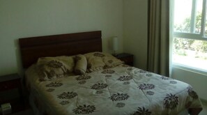 A neatly made bed with a floral bedspread, a wooden headboard, a bedside table with a lamp, and a window with a view of greenery. - 56.- HNA Capri (La Serena)