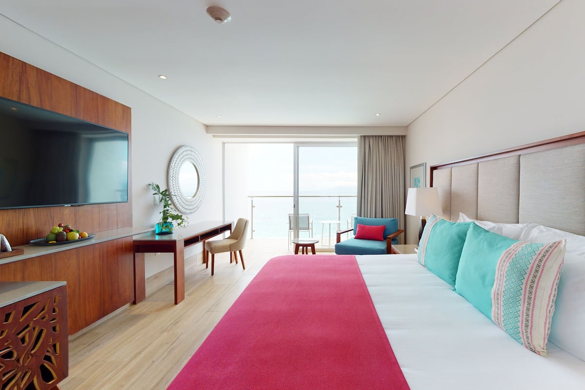 Une chambre d’hôtel dotée d’un grand lit, d’un bureau avec un téléviseur à écran plat, d’un coin repas avec une table et des chaises, et d’un balcon offrant une vue sur la mer.