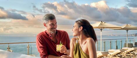 Una pareja sentada en una mesa con vista al mar, sillones de descanso y sombrillas al fondo.