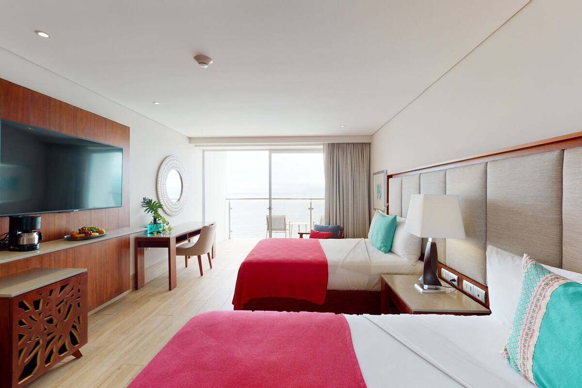 Une chambre d’hôtel équipée d’une télévision, d’un bureau avec une machine à café, de deux lits avec une literie rouge et blanche, d’une grande fenêtre offrant une vue et d’un balcon avec une chaise.