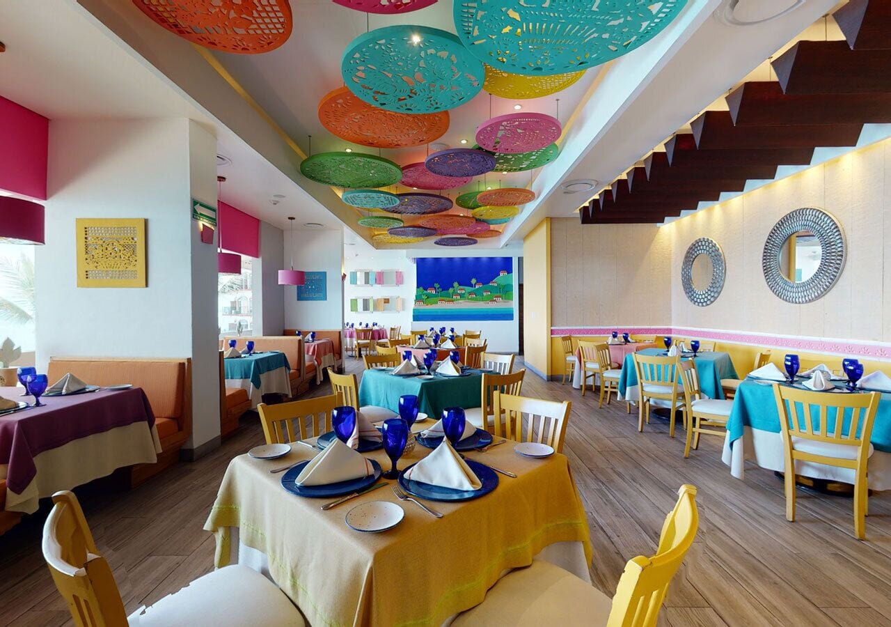 A restaurant with multiple tables set for dining, colorful hanging decorations, and a view of the sea in the background.