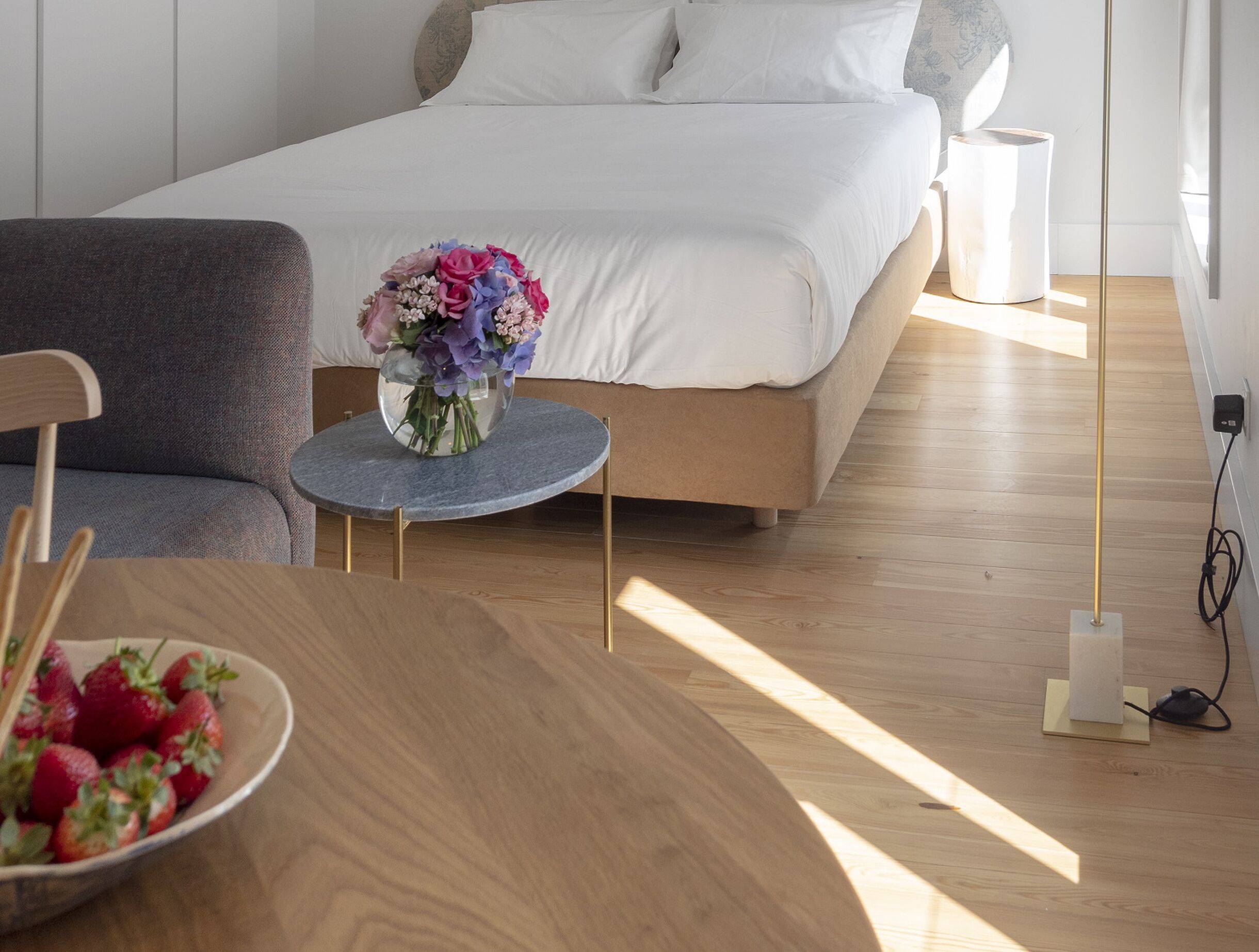 Une chambre à coucher avec un lit, une table ronde ornée d’un vase de fleurs, un bol de fraises et une lampe de chevet.