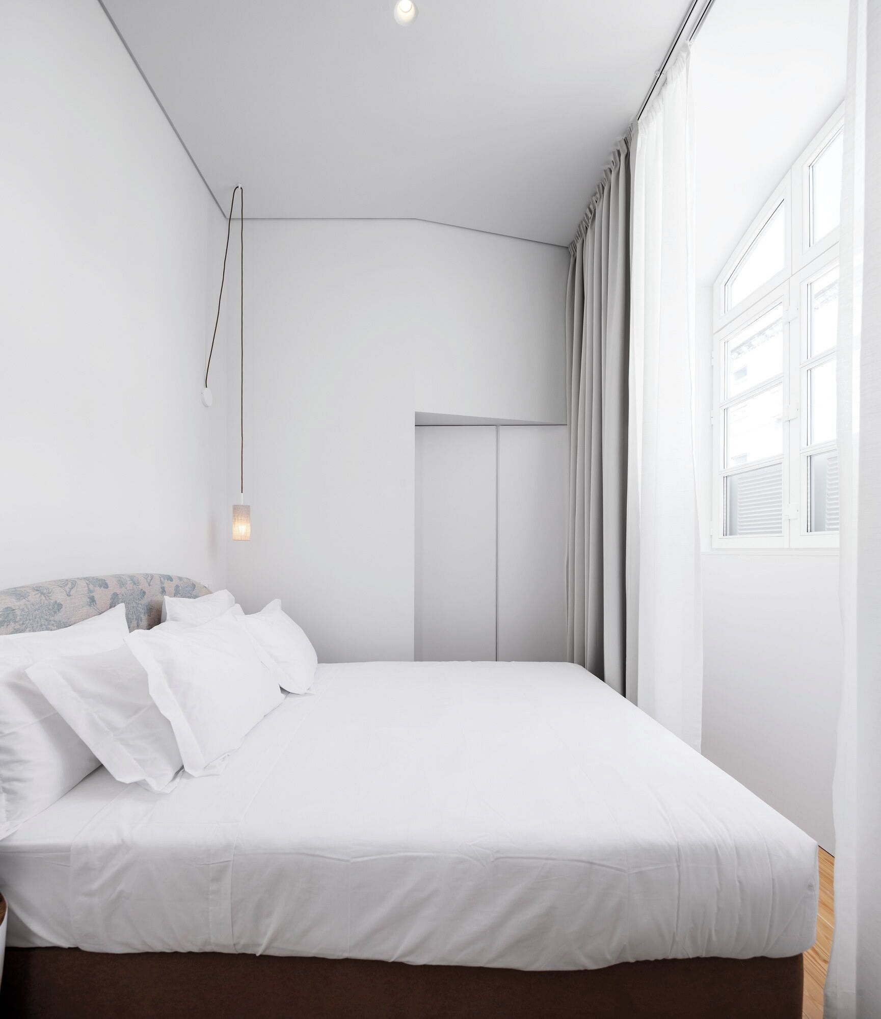 A modern bedroom with a large bed, a bedside table, a hanging light, and a window with curtains.