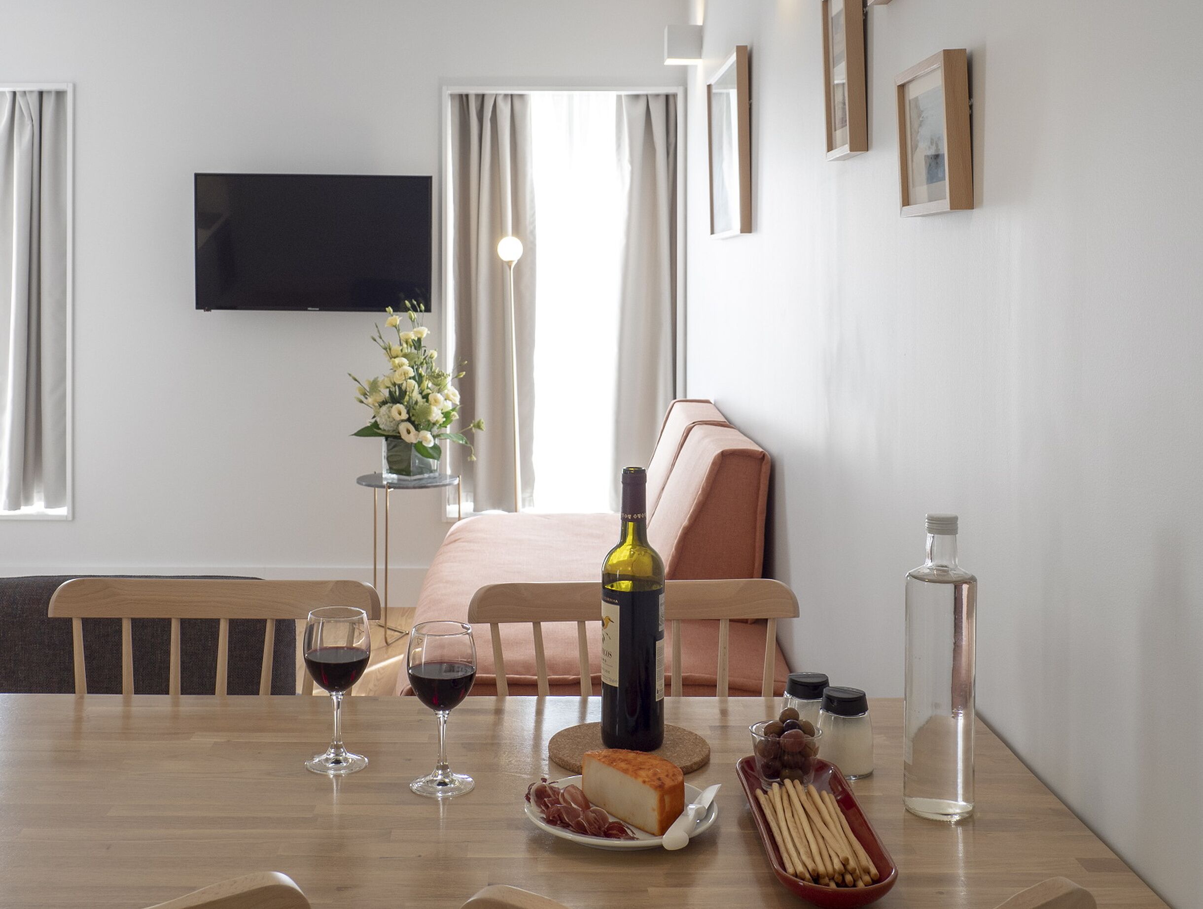 Camera d'albergo con un letto, un tavolo da pranzo con bicchieri da vino, una bottiglia di vino, un piatto di formaggio e un vaso di fiori.