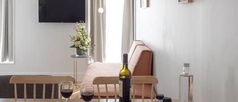 A hotel room with a bed, a dining table with wine glasses, a bottle of wine, a plate of cheese, and a vase of flowers.