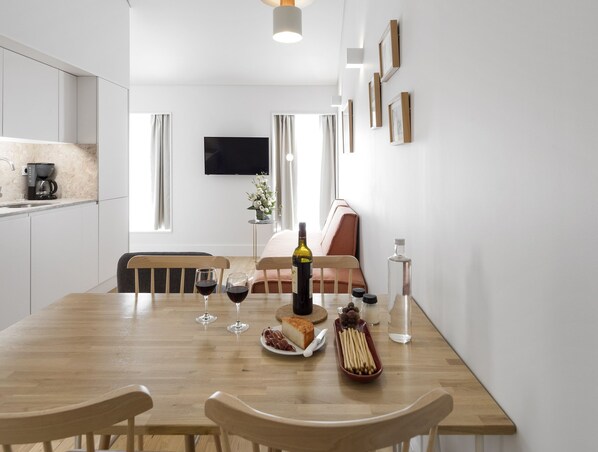 A modern kitchen with a dining table set for two, a coffee machine, and a flat-screen TV mounted on the wall.