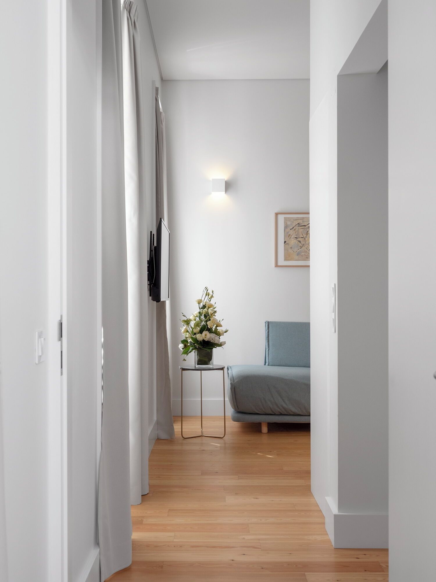 A narrow hallway with a wooden floor, a small table with a vase of flowers, a grey sofa, and a framed picture on the wall.