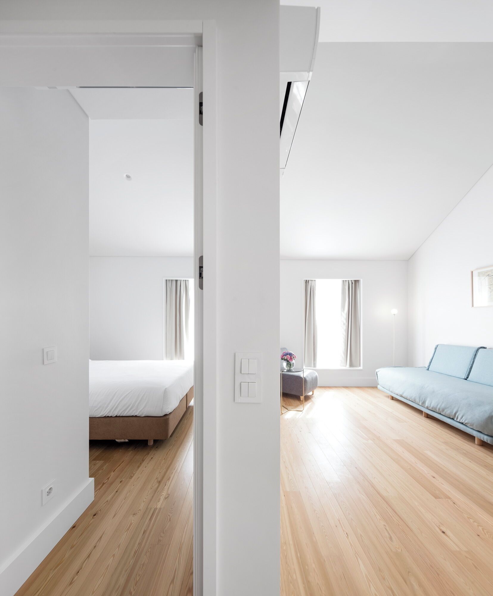 A modern, minimalist bedroom with a wooden floor, a bed, a blue sofa, and a small table with a vase.
