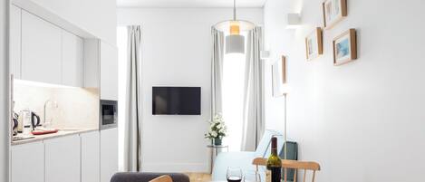 A modern kitchen with white cabinets, a dining table with chairs, a TV mounted on the wall, and a wooden floor.