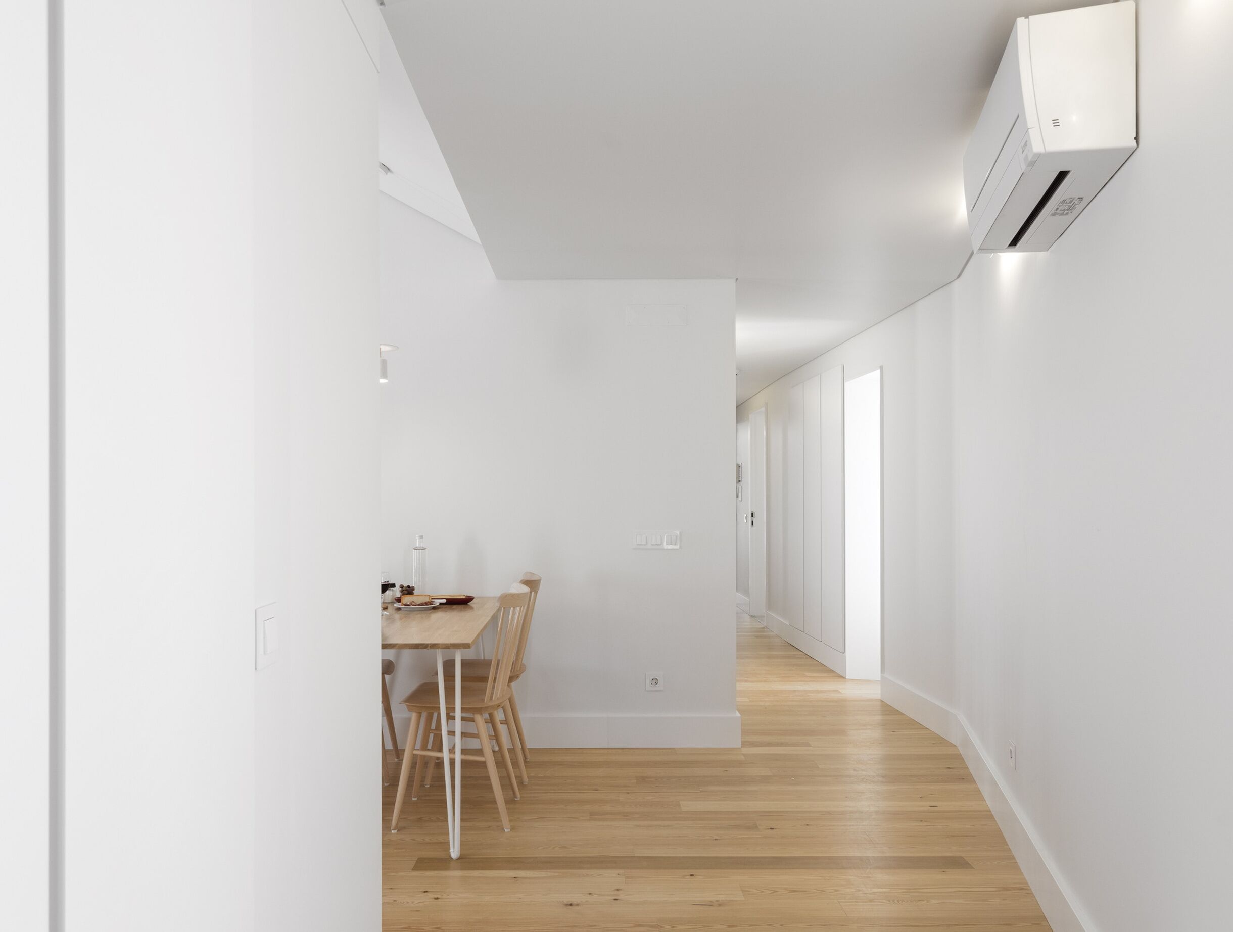 A narrow hallway with a dining area featuring a wooden table and chairs, white walls, and a wooden floor.