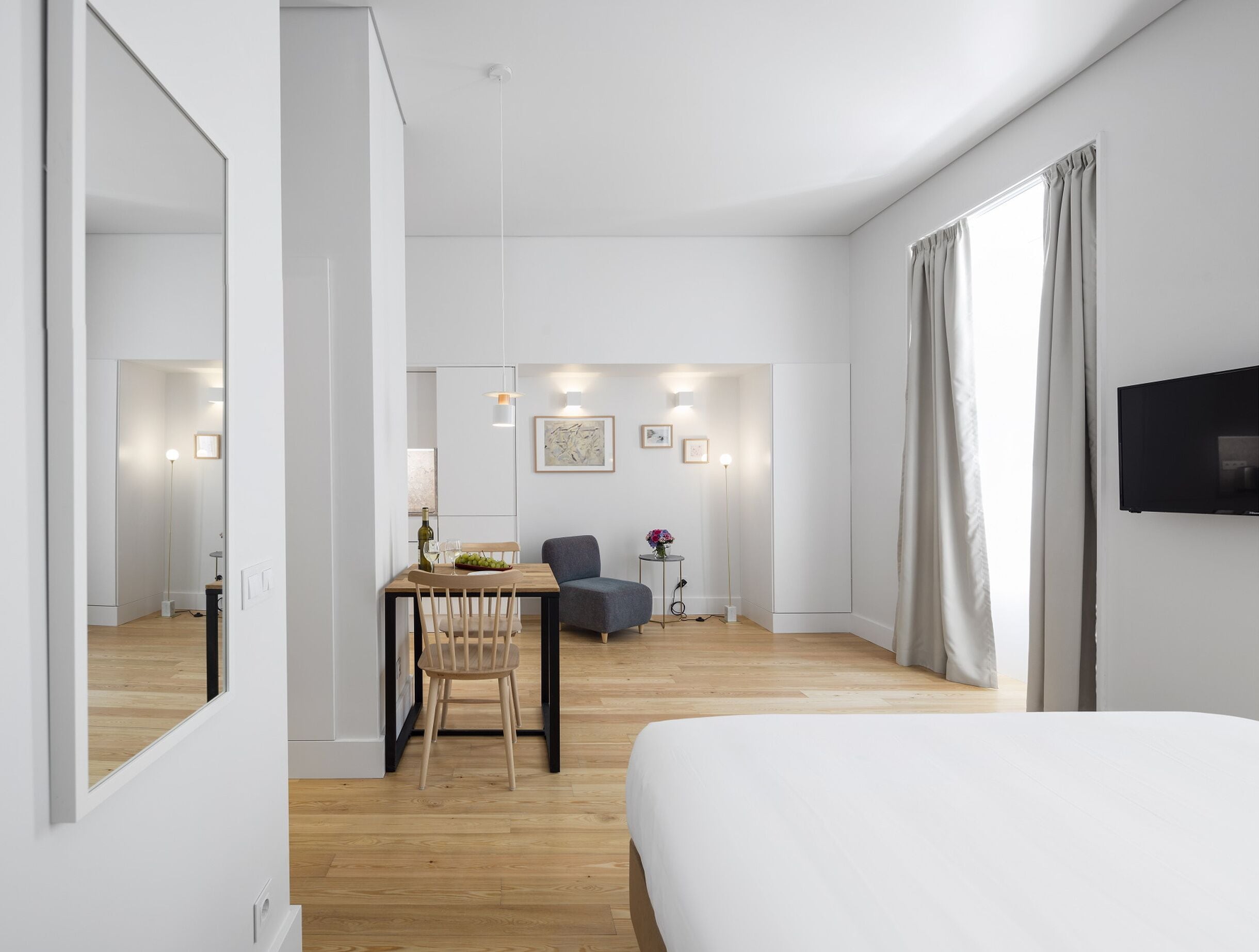 A modern hotel room with a large bed, a dining area with a table and chairs, a TV mounted on the wall, and a large window with curtains.