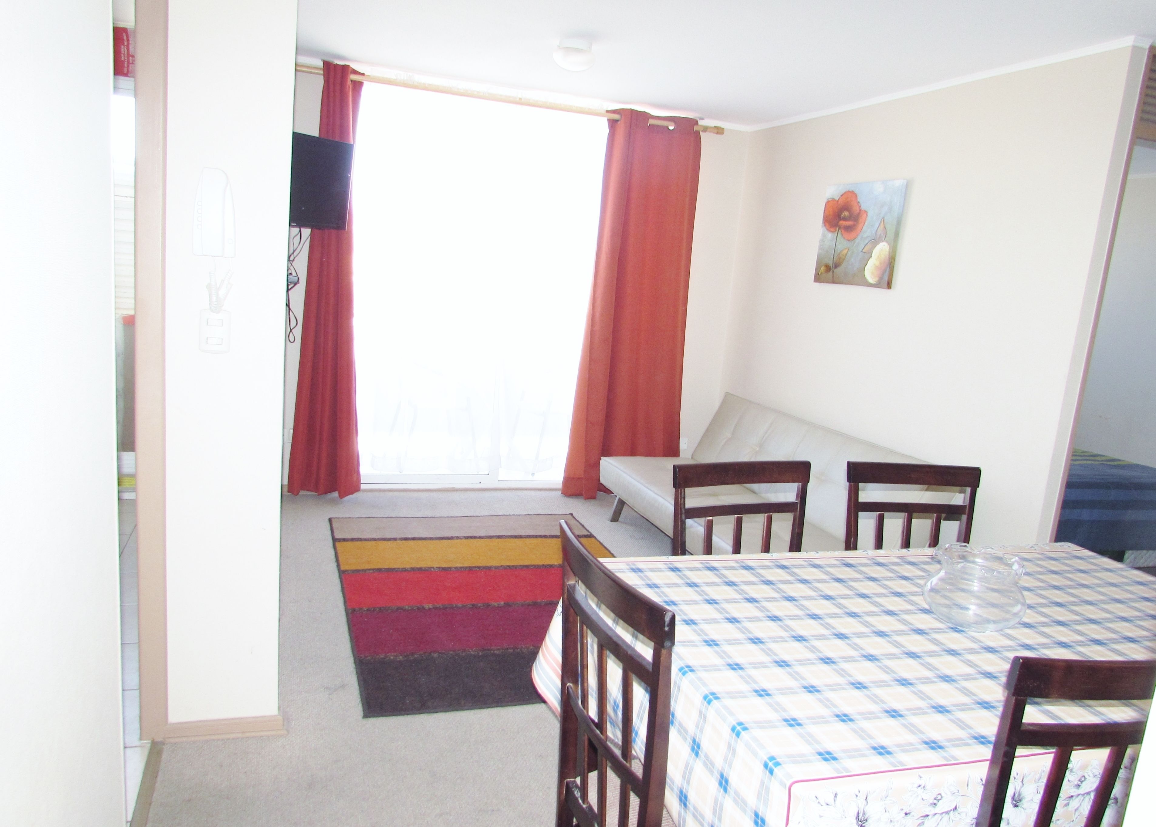 Una habitación con una mesa a cuadros, sillas de madera, un sofá, una cortina roja y una ventana con vistas.