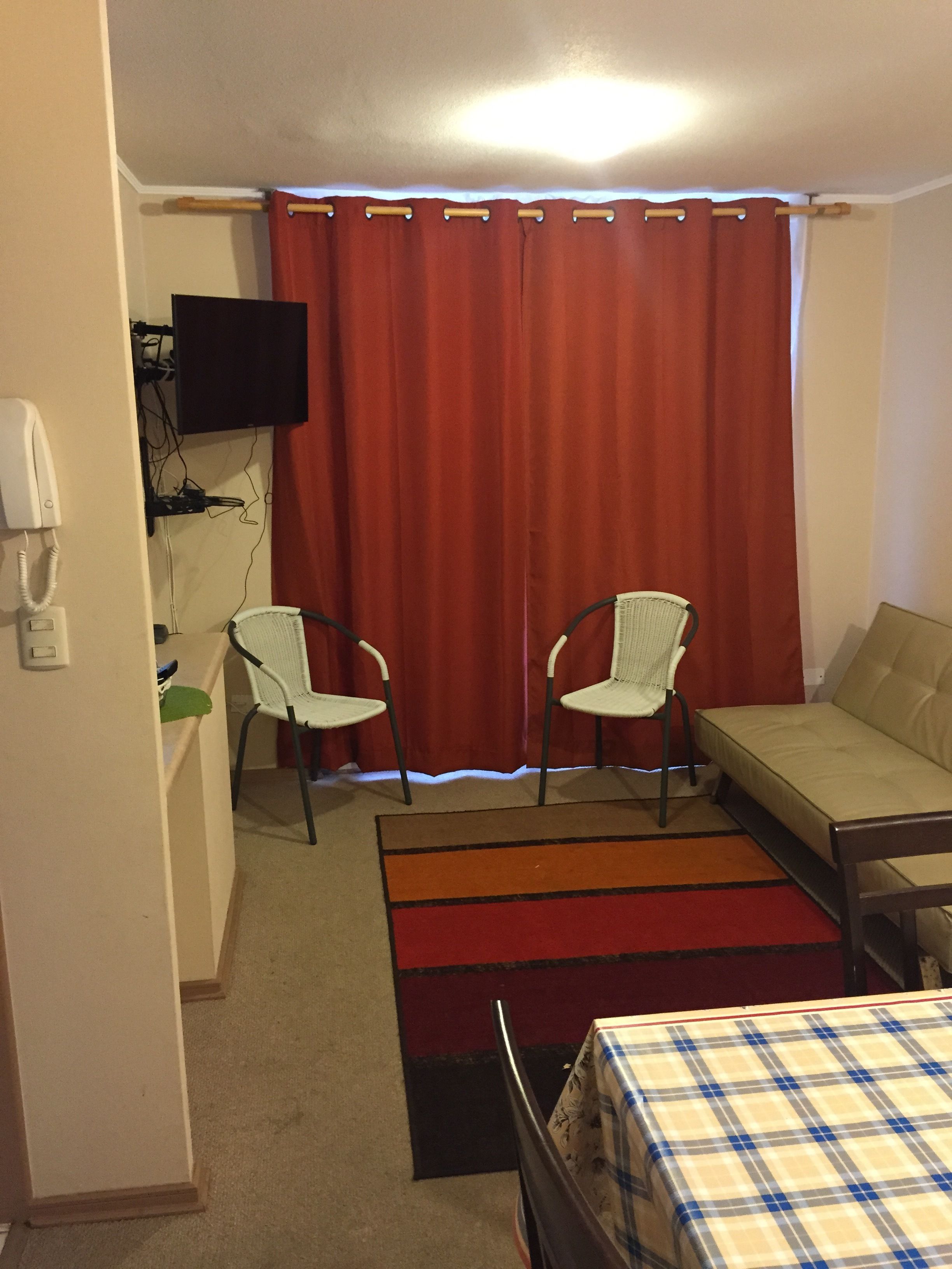 Una habitación con dos sillas blancas, un sofá, un televisor de pantalla plana montado en la pared, un teléfono y una cortina roja.