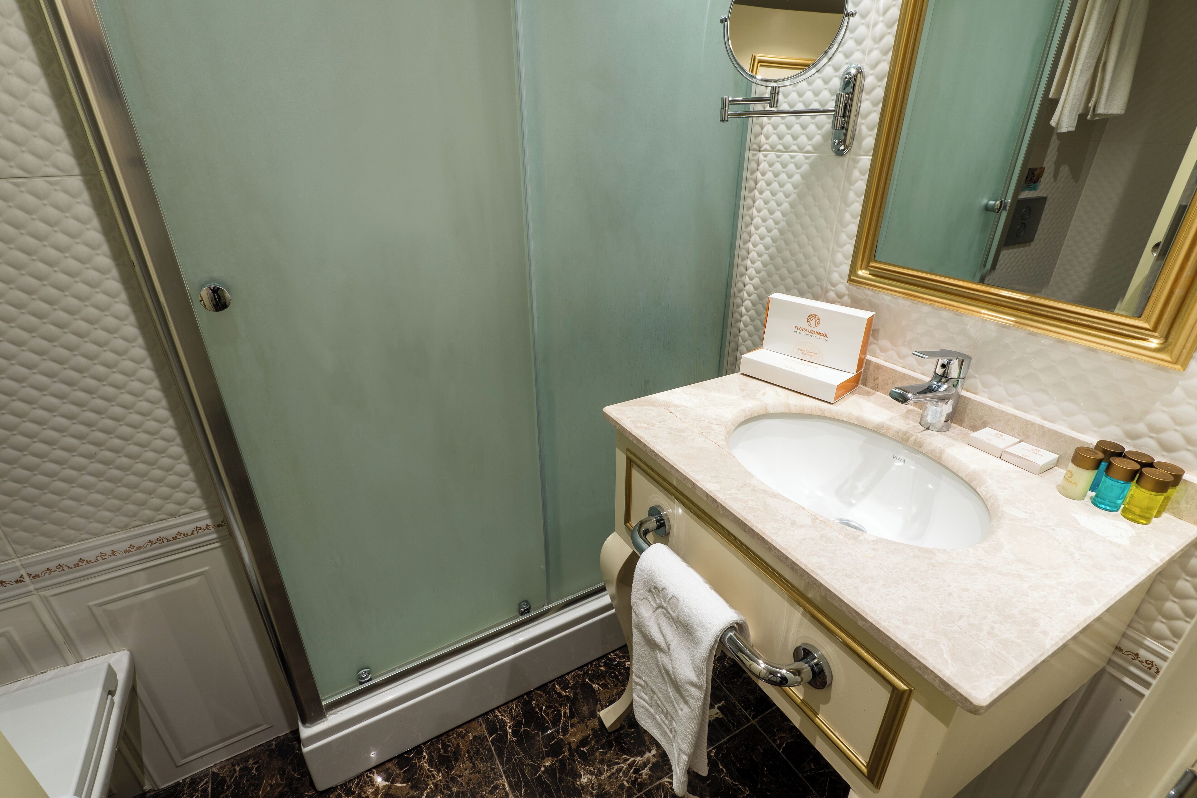 A bathroom with a glass shower enclosure, a sink with a marble countertop, and a mirror with a gold frame.