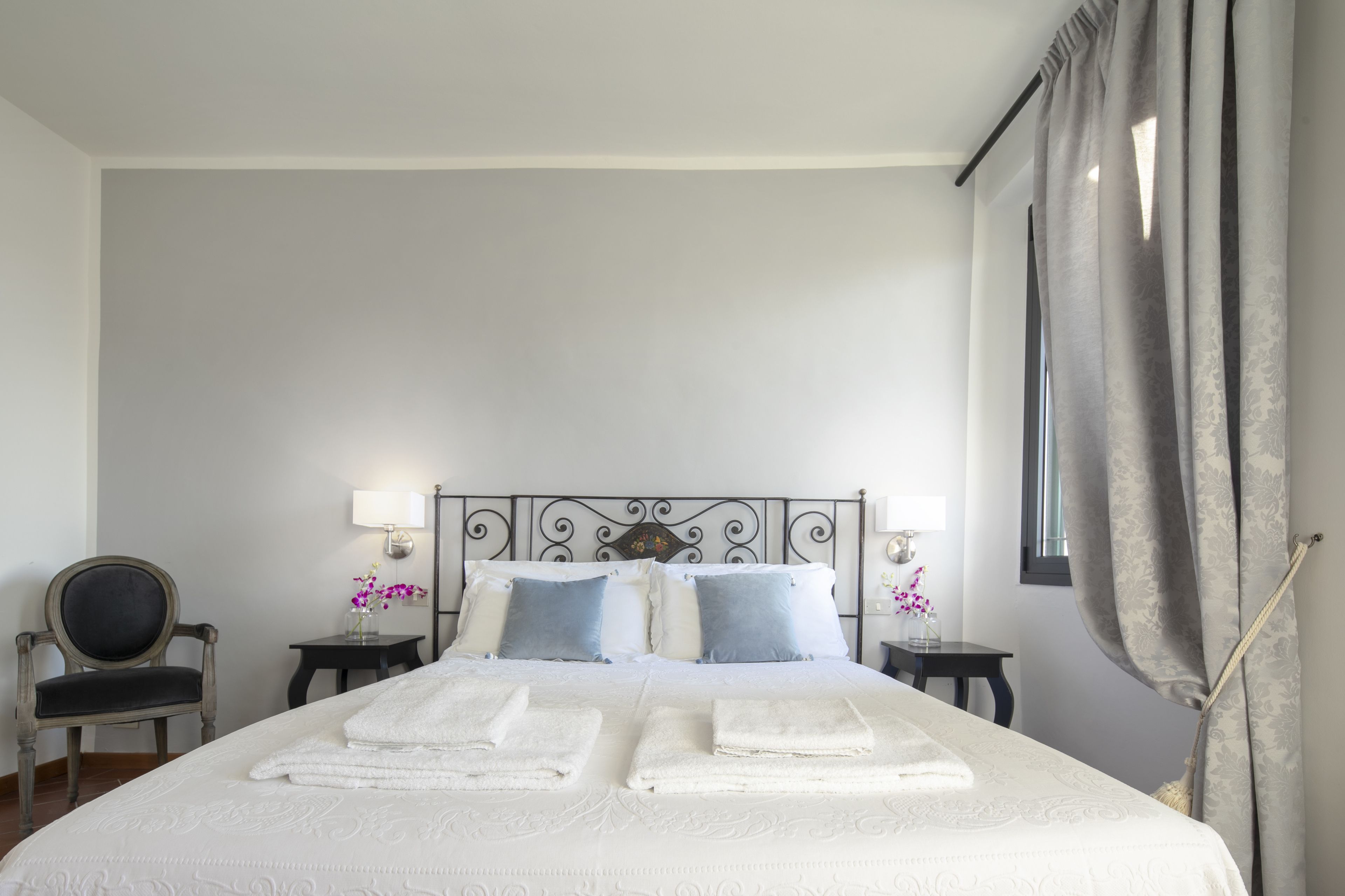 A neatly made bed with folded white towels, a metal headboard, two bedside tables with lamps, a chair, and a window with curtains.