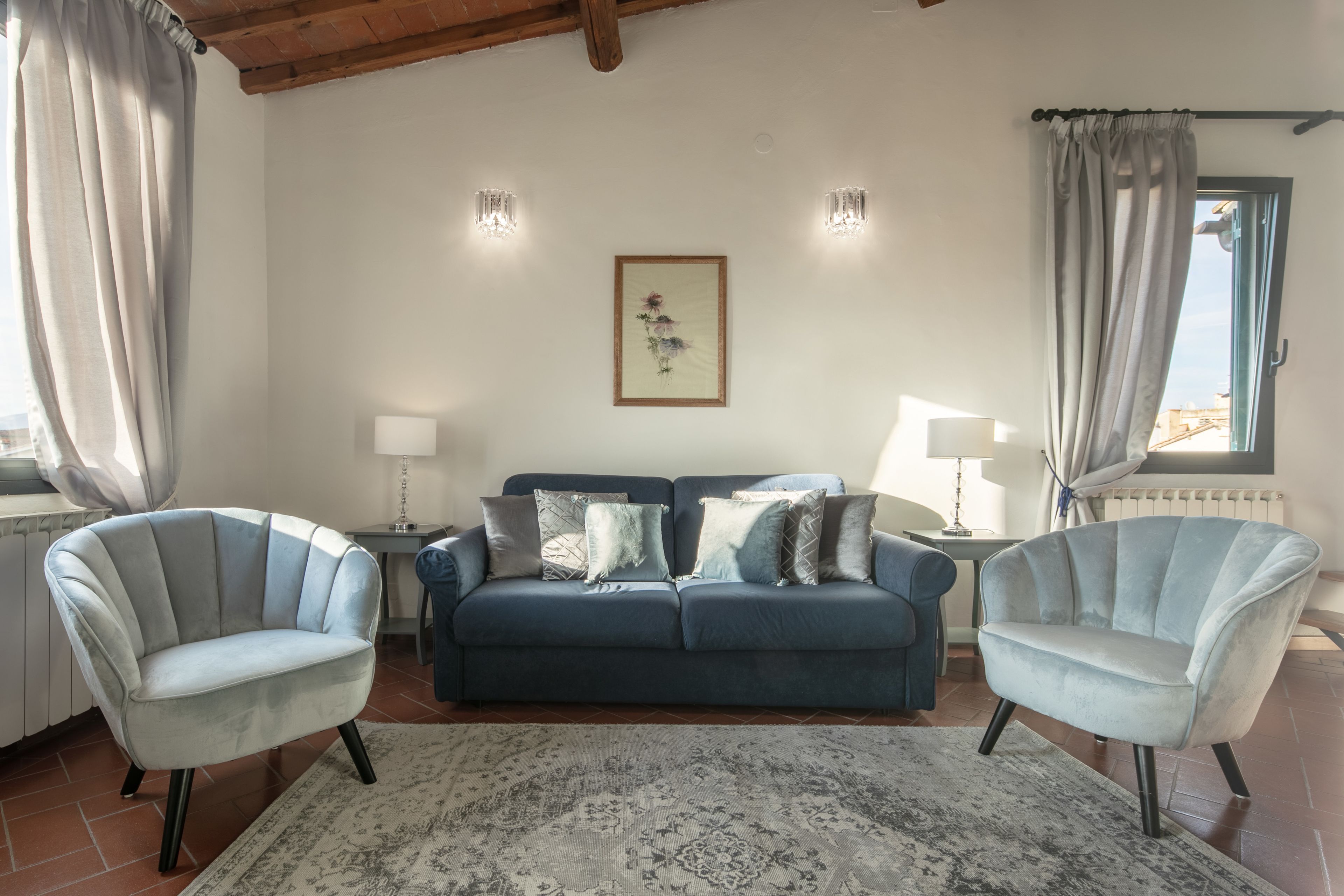 Un salon avec un canapé bleu, deux fauteuils, un tapis à motifs et une œuvre d’art encadrée accrochée au mur.