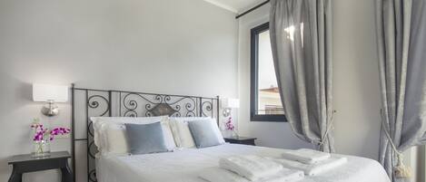 A neatly made bed with white linens and a decorative headboard, a bedside table with a lamp, and a window with grey curtains.