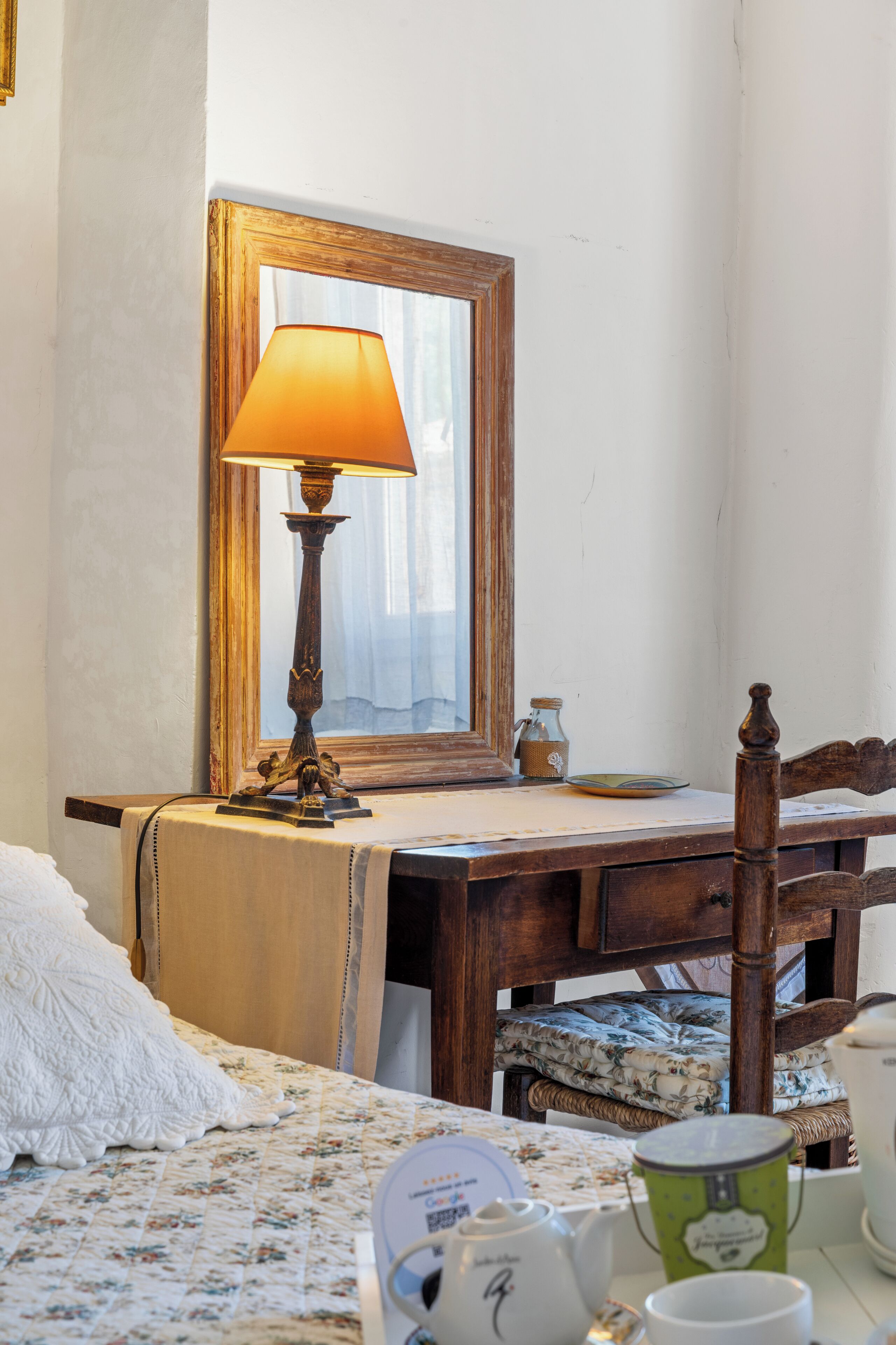 A room with a wooden desk, a lamp, a mirror, a bed with a floral bedspread, and a wicker chair.