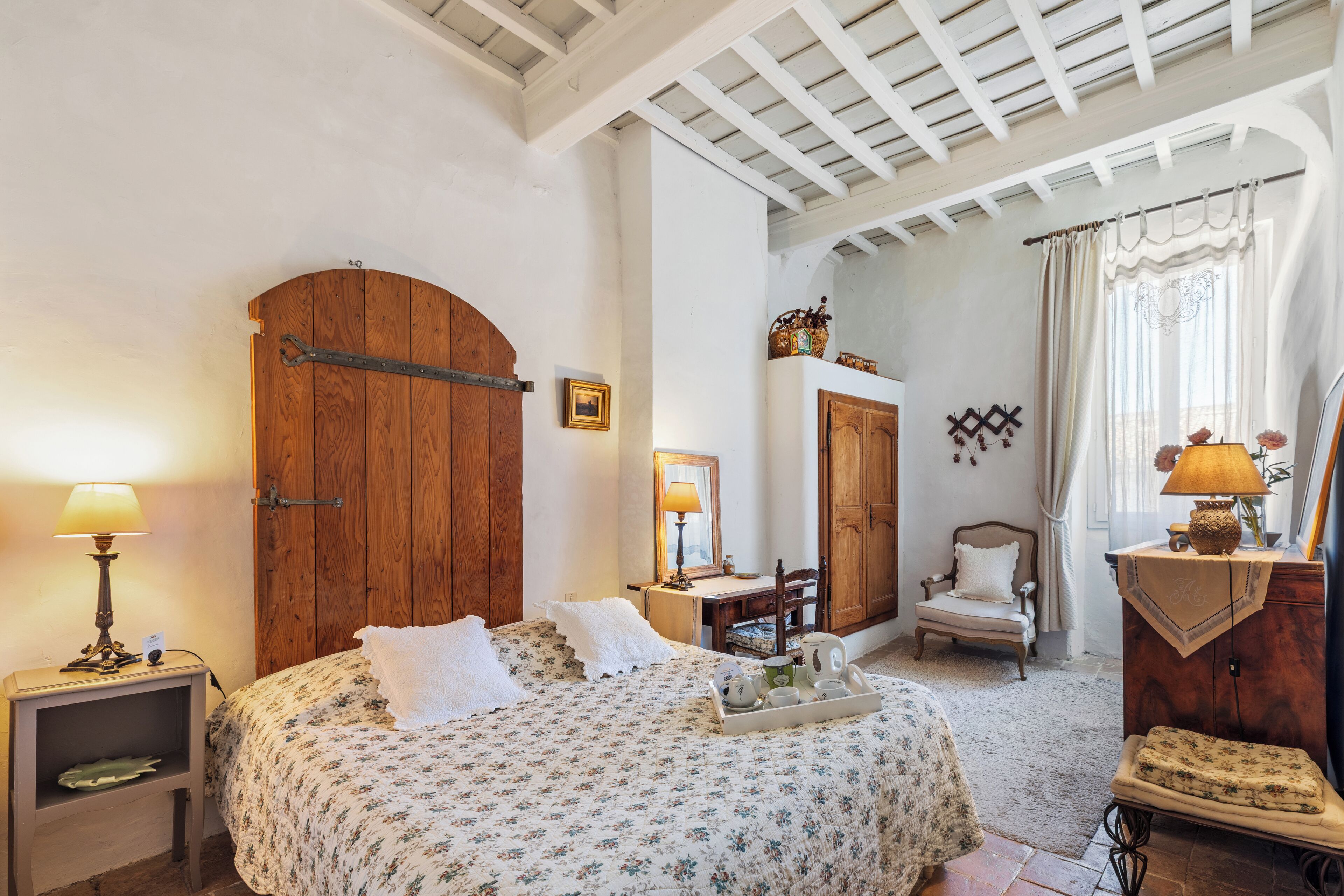 Une chambre avec une porte en bois, un lit avec une literie à motifs floraux, une table de chevet avec une lampe, une chaise et une commode.