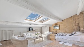 Une chambre spacieuse aménagée en mezzanine, avec un plafond en pente, des fenêtres de toit, un grand lit, un canapé blanc et des fauteuils en osier.