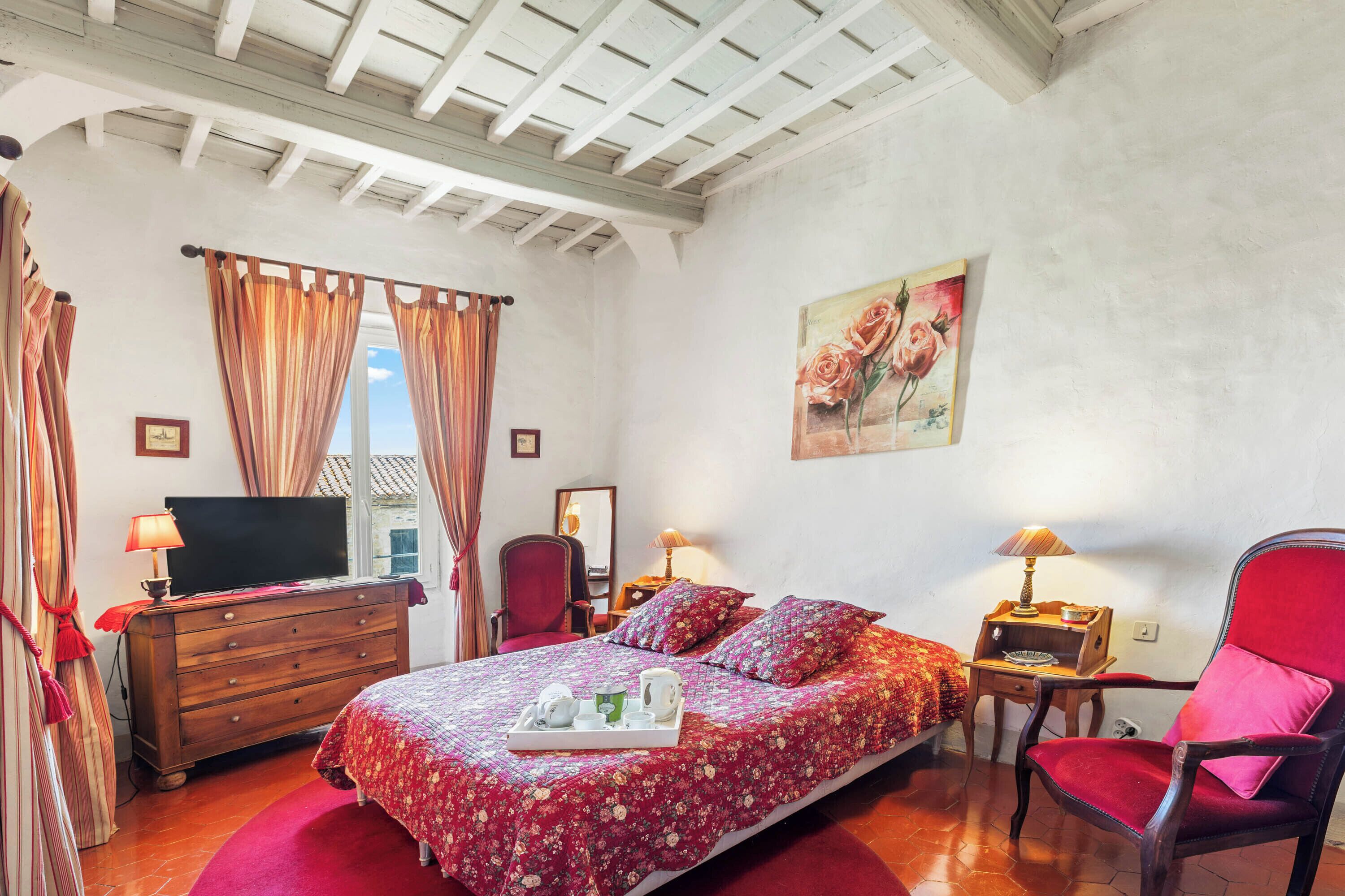 A bedroom with a bed, a TV, a dresser, a chair, and a table with a lamp.