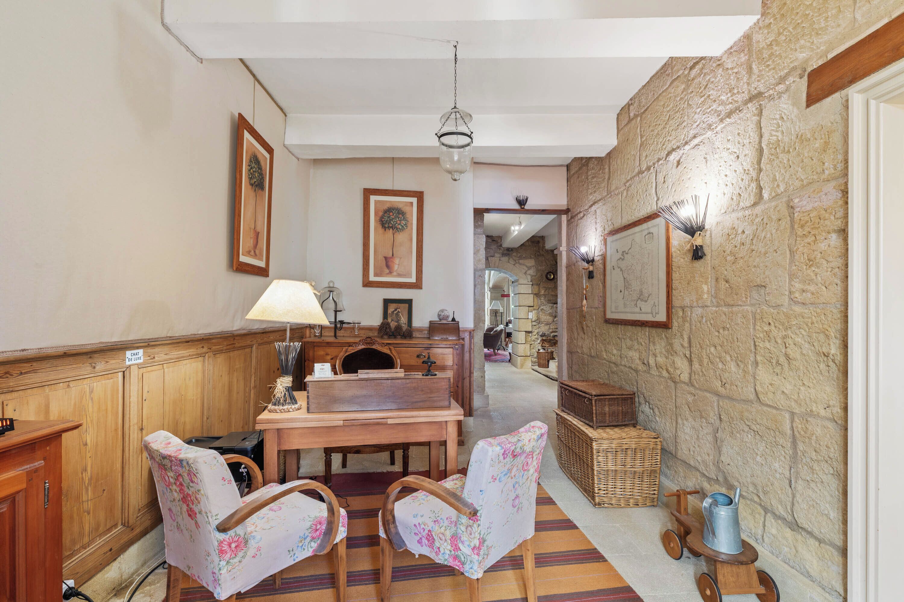 A cozy room with a wooden table, two floral chairs, a lamp, and a stone wall.