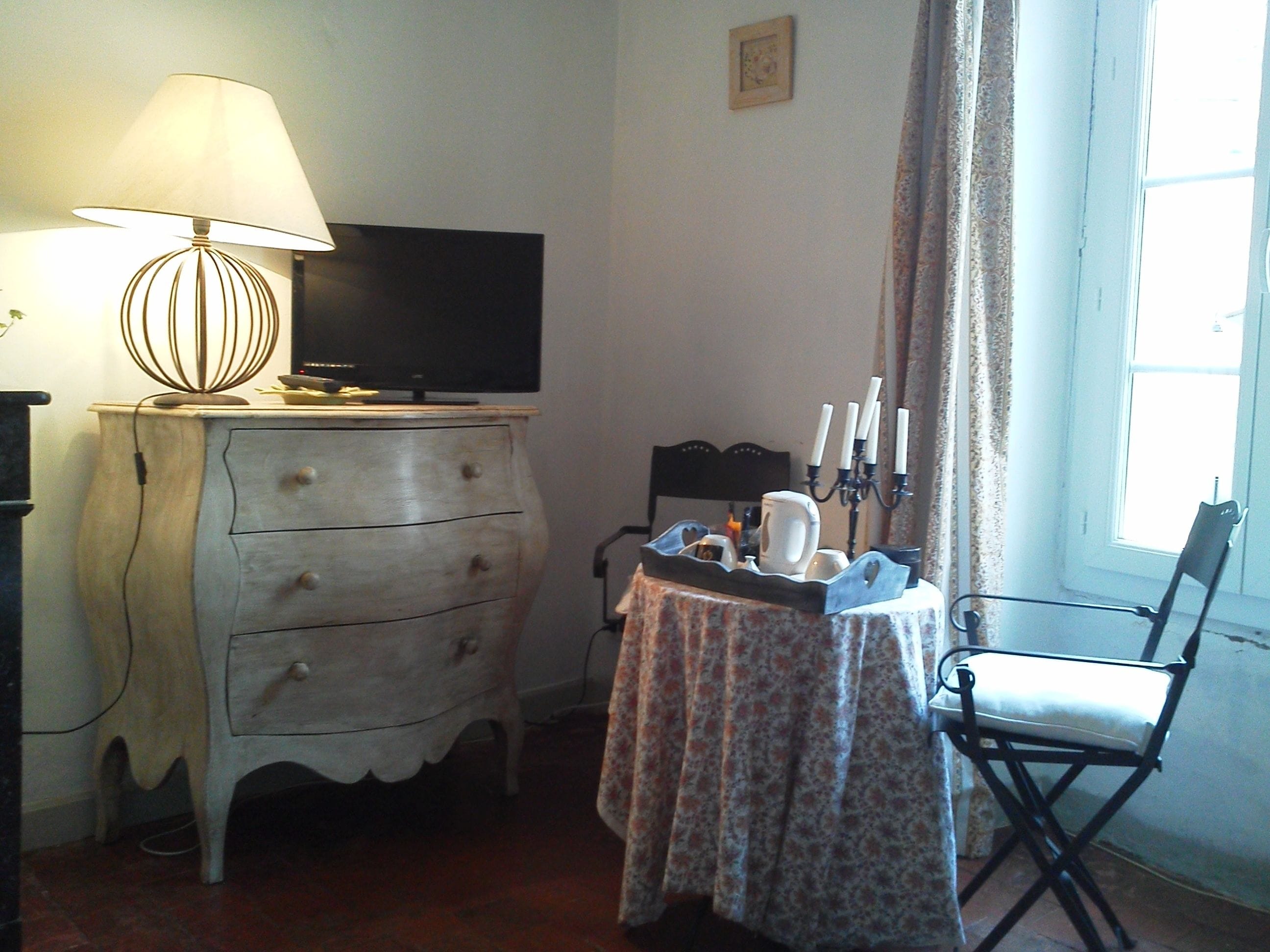 Une pièce avec une télévision posée sur une commode, une table recouverte d’une nappe à motifs floraux, une chaise et une fenêtre avec des rideaux.