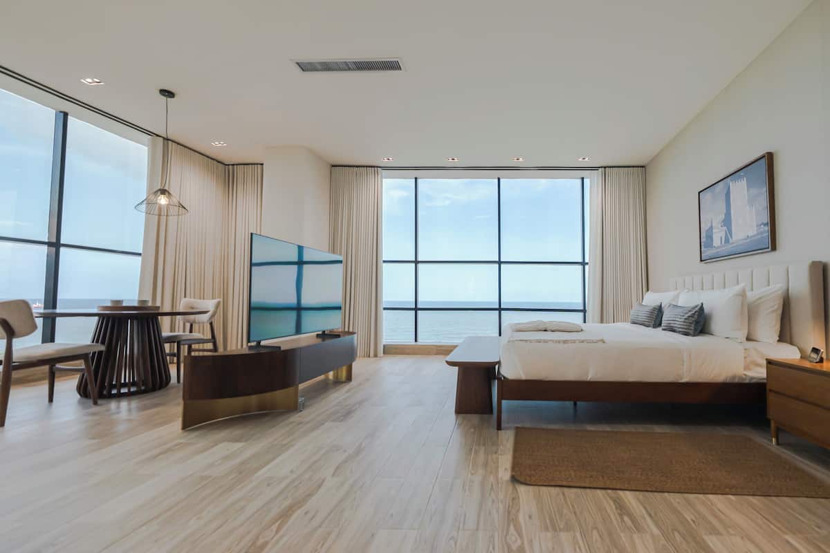 A modern hotel room with a large bed, a desk, a TV, and a view of the sea.