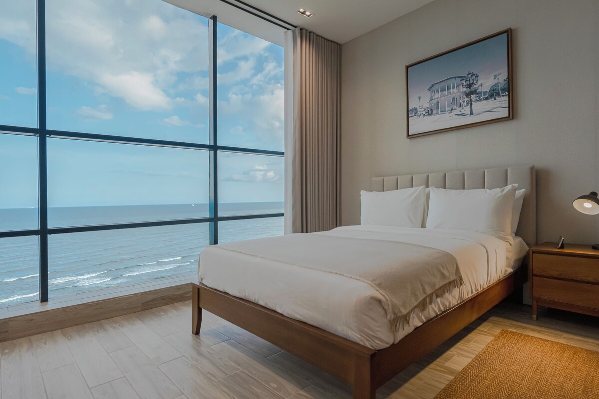 Un dormitorio con un amplio ventanal que ofrece vista al océano, una cama con ropa blanca, una mesita de noche con lámpara y un cuadro en la pared.