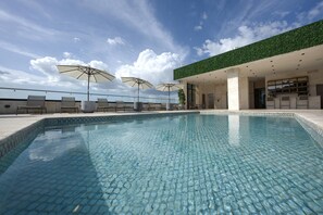 Una piscina con sillas de descanso y sombrillas, rodeada por un edificio con techo verde y cielo despejado.