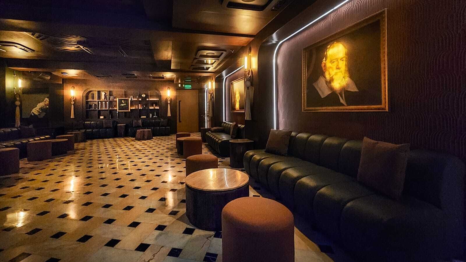 A dimly lit lounge area with a patterned floor, leather seating, and framed artwork on the walls.
