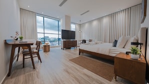 A modern hotel room with a large bed, a desk with a chair, a TV, and a view of the city through large windows. - Mauad Hotel Boutique (Santo Domingo)