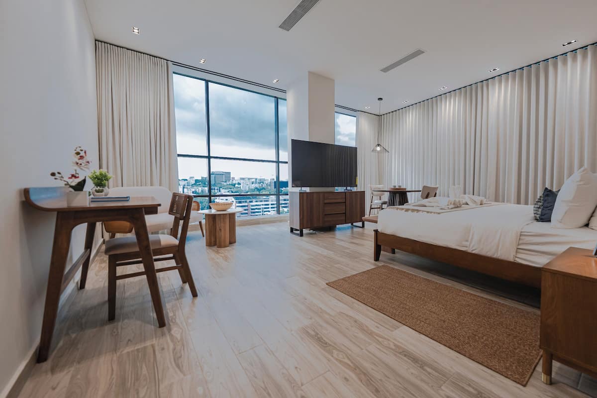 A modern hotel room with a large bed, a desk with a chair, a TV, and a view of the city through large windows.