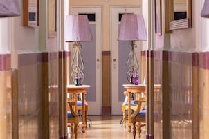 A hotel hallway with mirrored walls, two lamps, and wooden chairs with blue cushions.