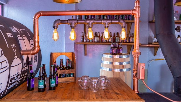Un espace bar avec des tuyaux en cuivre, des ampoules apparentes et des bouteilles exposées sur des étagères.