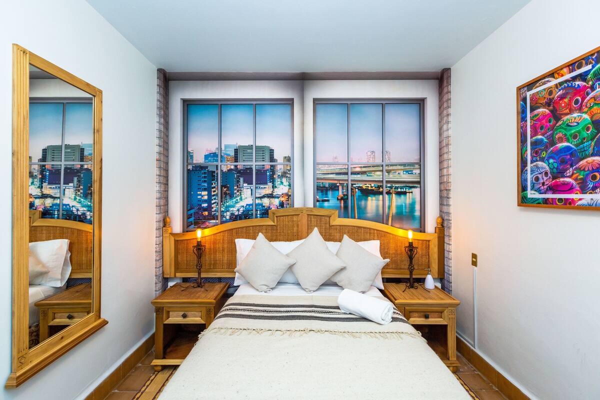 Une chambre d’hôtel avec un grand lit, deux tables de chevet, un miroir et une vue sur la ville par les fenêtres.