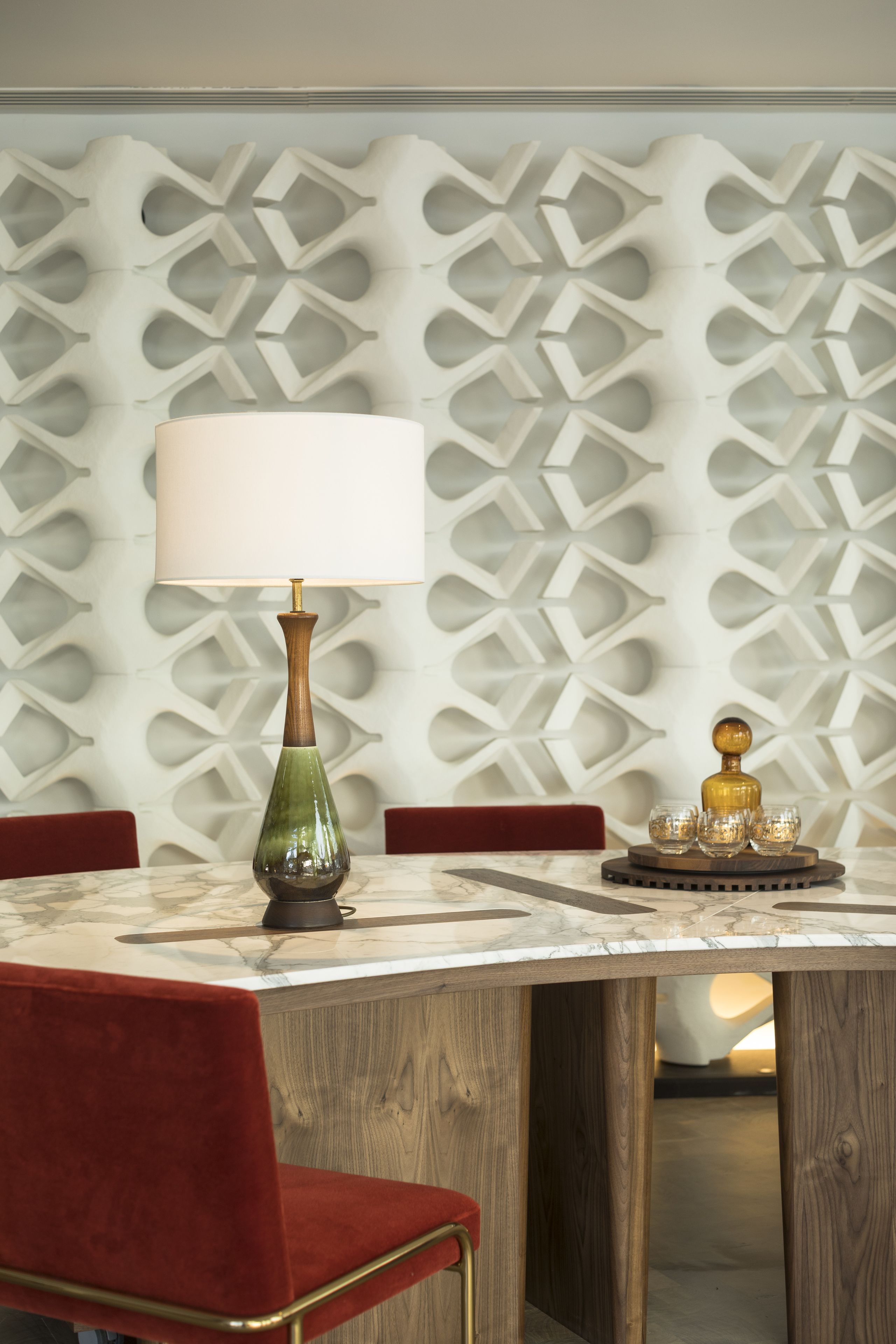 A dining area with a marble table, red upholstered chairs, a decorative wall, and a table lamp.