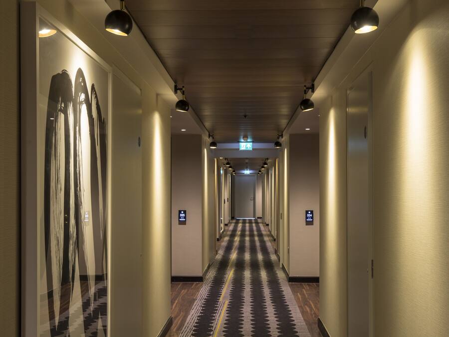 A hotel hallway with a patterned carpet, wall-mounted lights, and doors with signage.