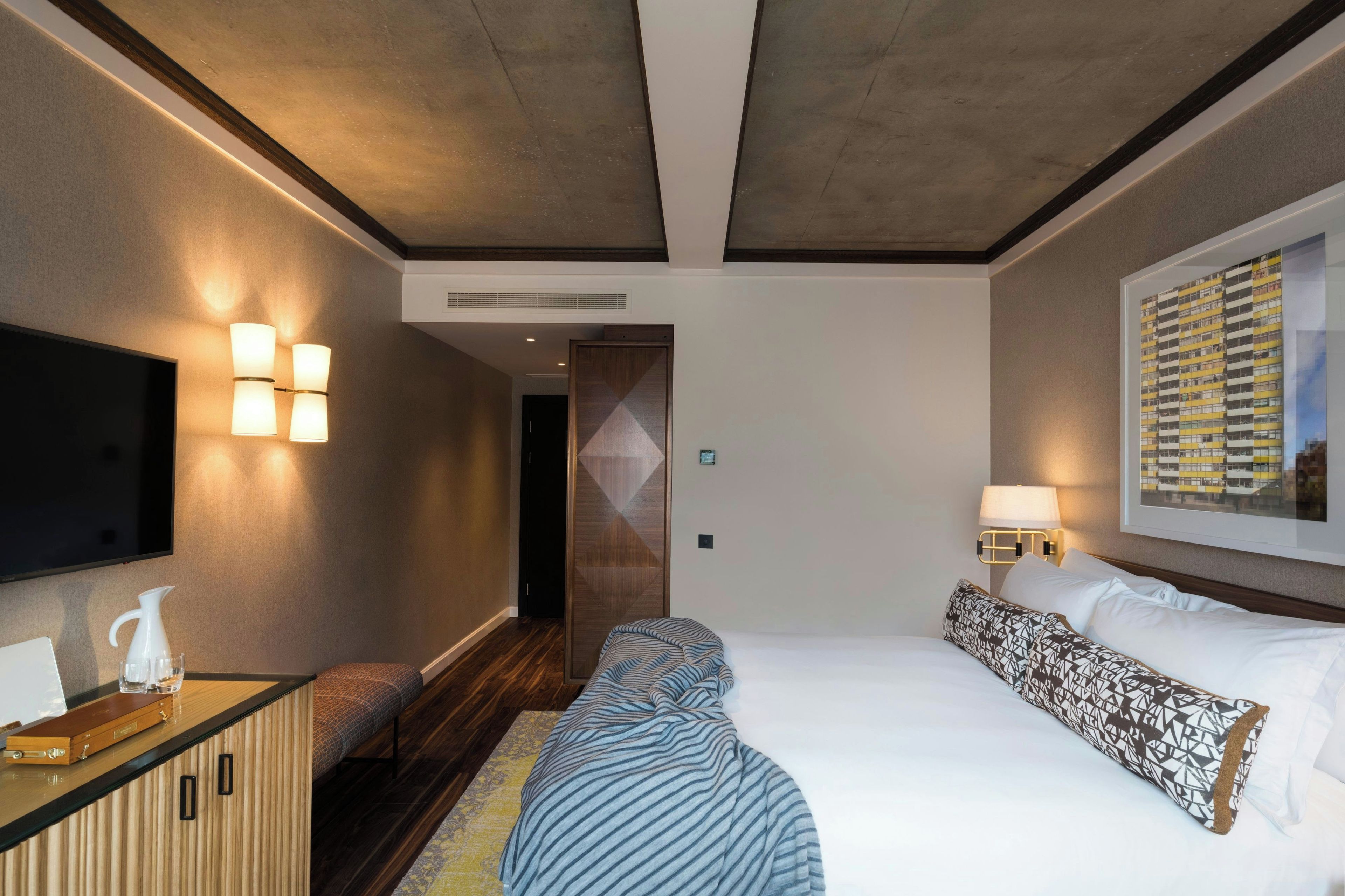 A hotel room with a bed, a TV, a desk, and a wall-mounted lamp.