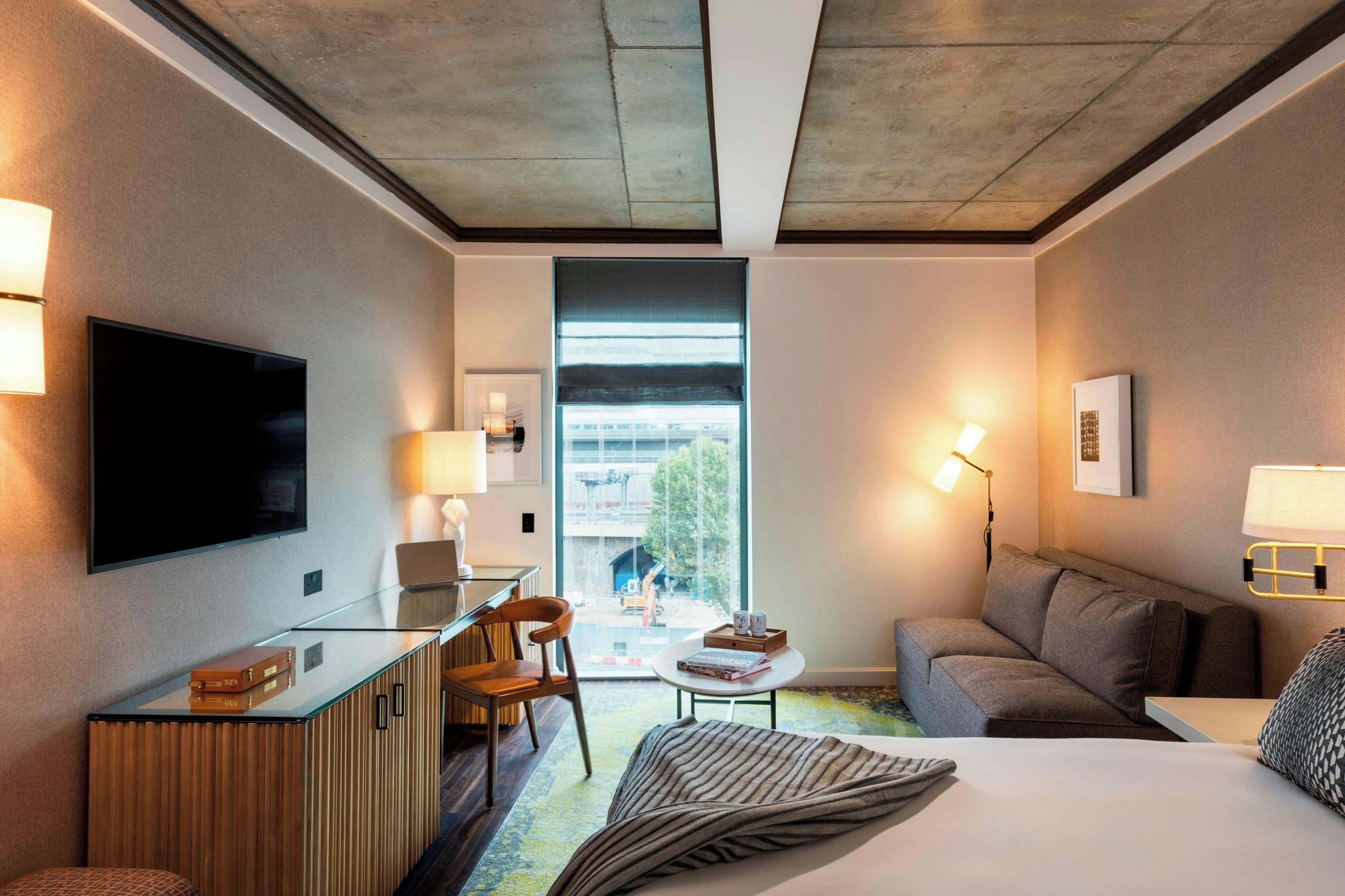 A modern hotel room with a flat-screen TV, a desk, a comfortable sofa, a bed, and a view of the outside.
