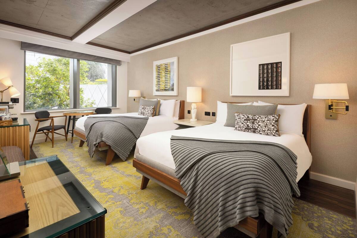 A hotel room with two beds, a desk, and a chair. There are two wall-mounted lamps and a window with a view of greenery.