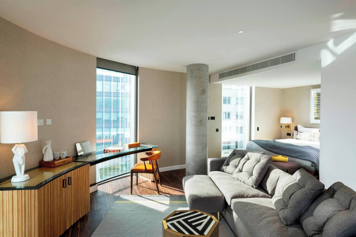 A modern hotel room with a large window, a balcony, a sofa, a desk, and a bed.