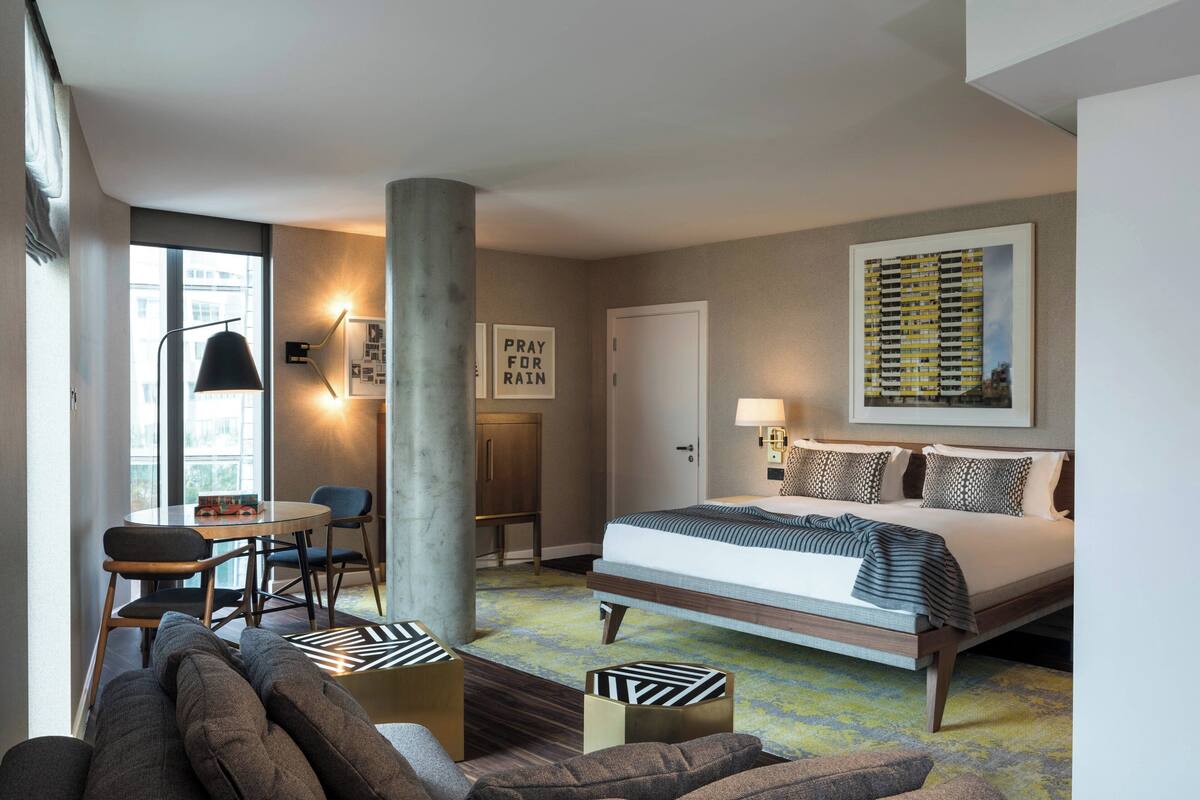 A modern hotel room with a large bed, a desk, and a sofa. There is a window with a view of buildings outside.