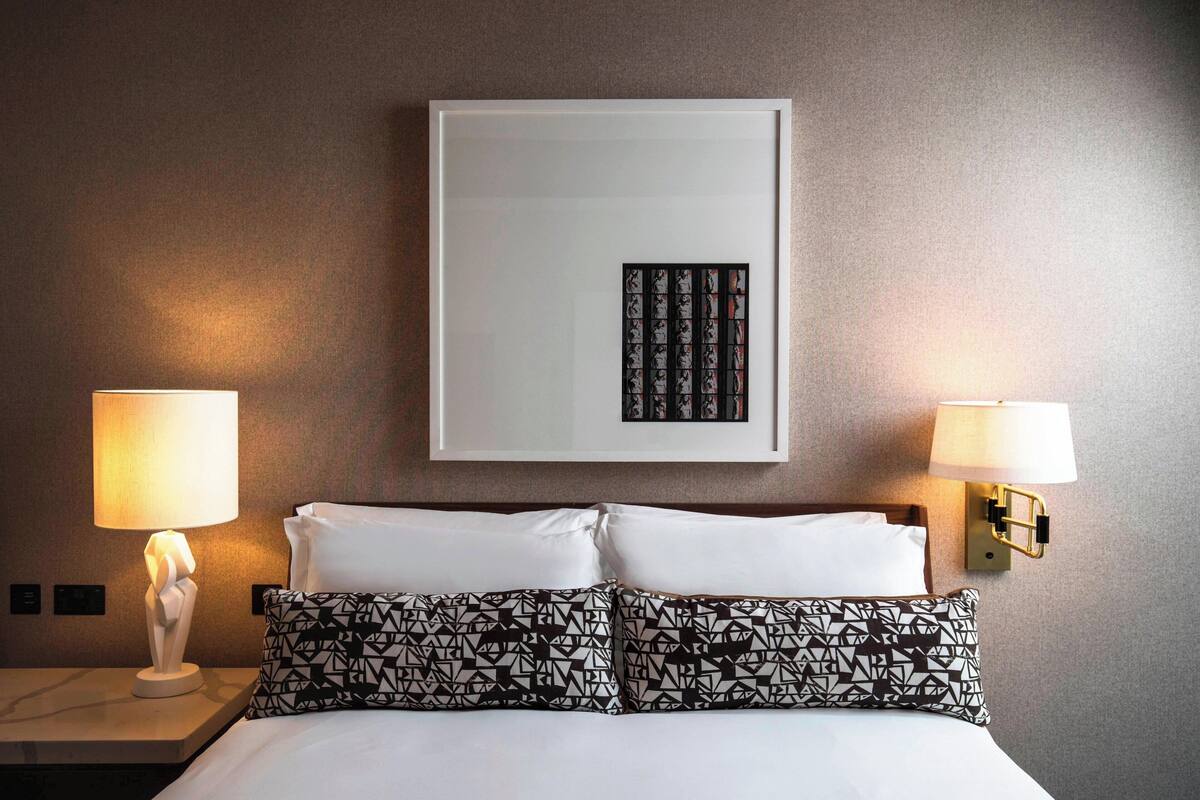 A hotel room with a bed, two bedside lamps, a framed artwork on the wall, and a white nightstand.