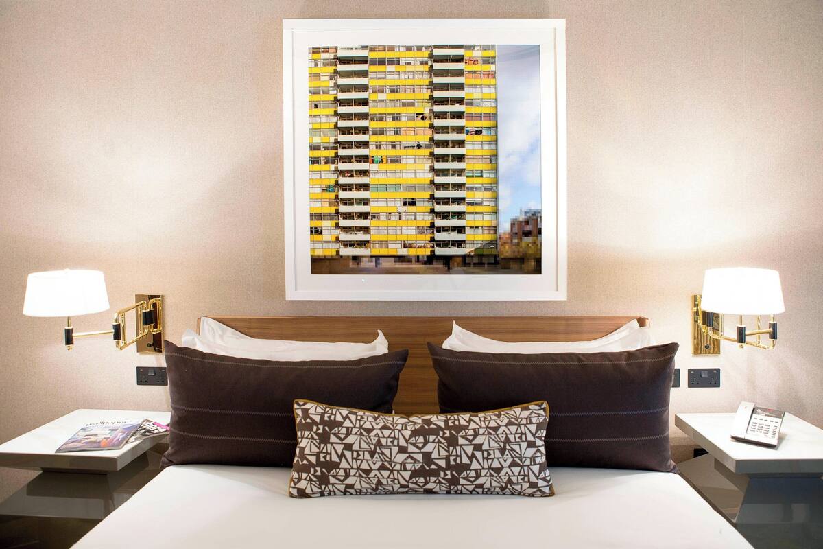 A hotel room with a large bed, two nightstands, a wall-mounted lamp, a framed cityscape picture, and a telephone on the nightstand.
