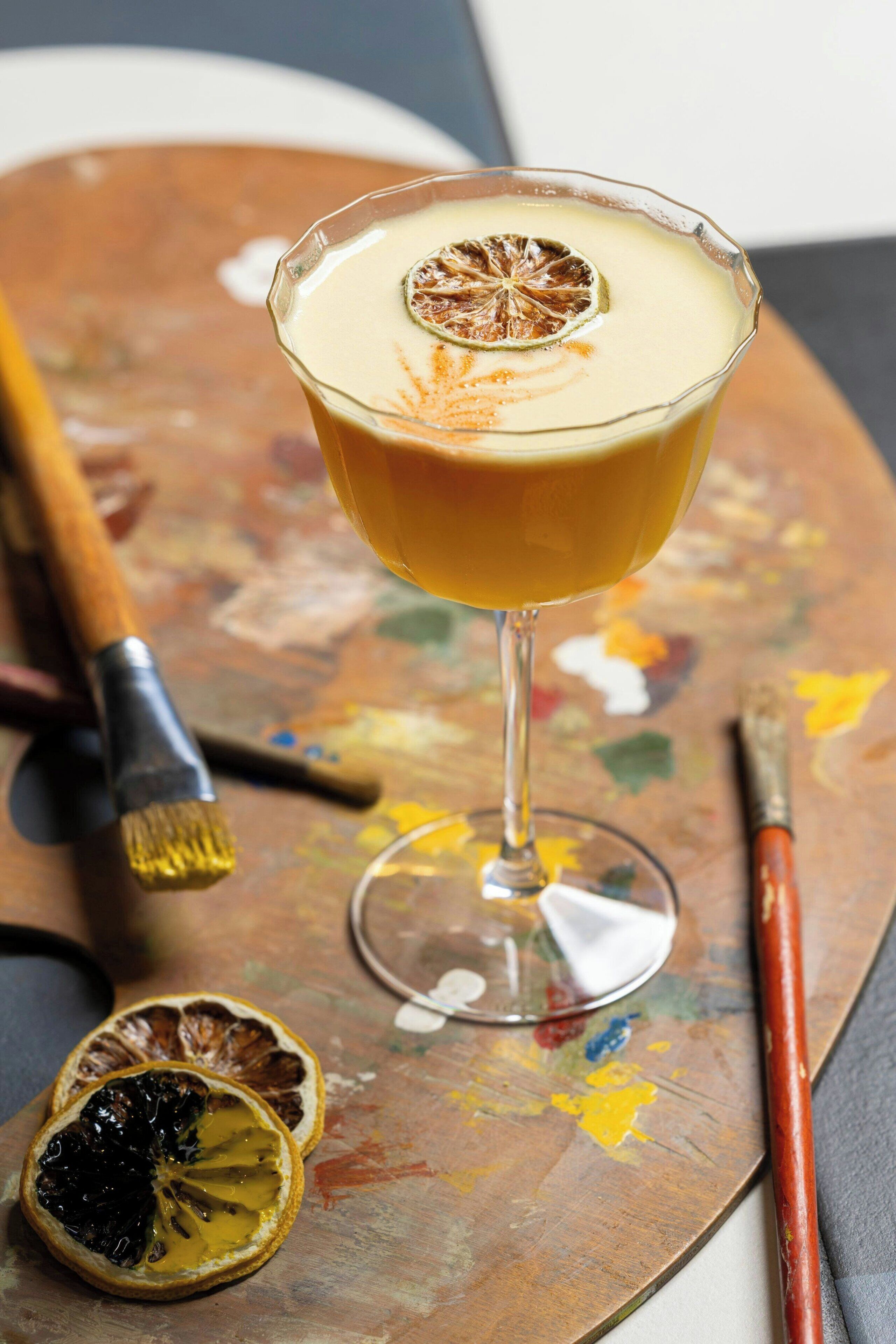 A cocktail glass with a dried orange slice garnish on a palette with paint and a brush.
