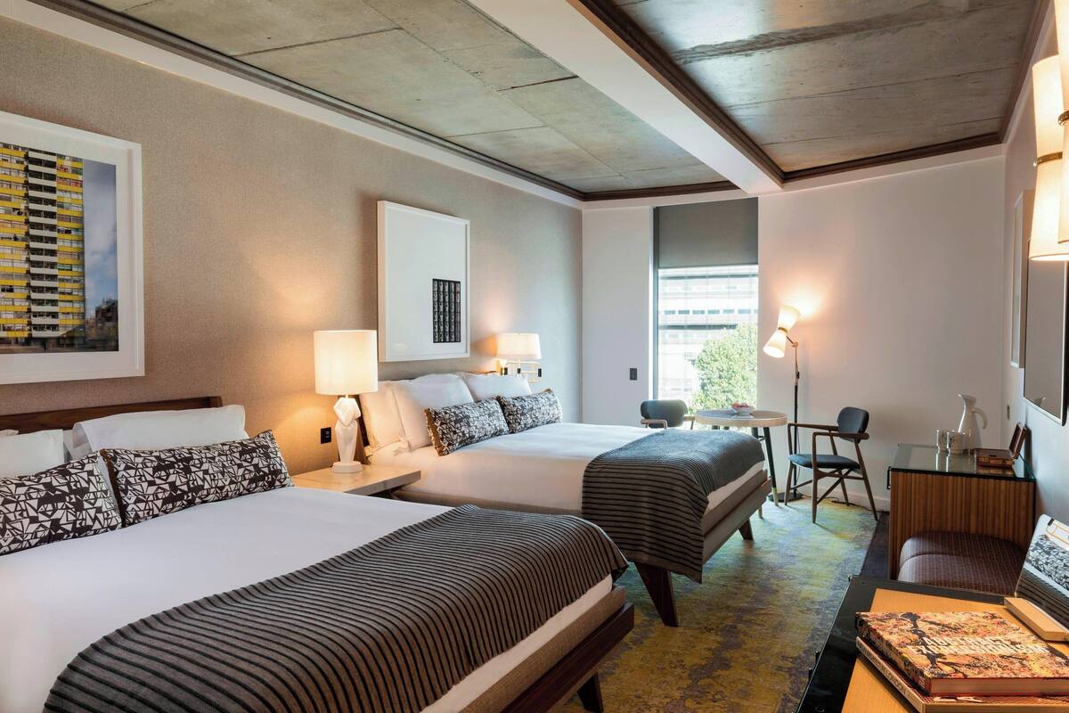 A hotel room with two beds, a desk, a chair, and a small table with a book.