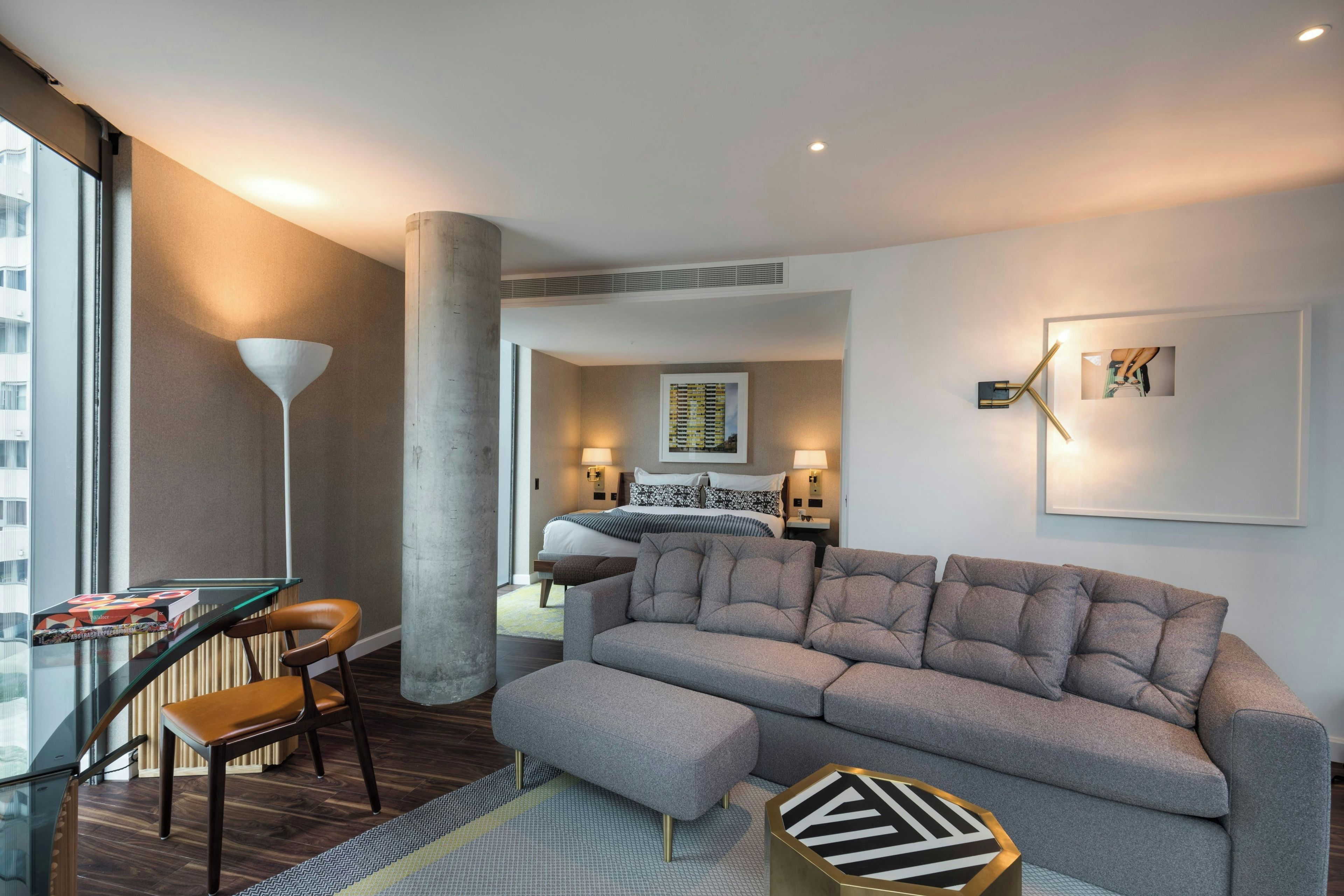 Una habitación de hotel moderna con un sofá gris, una mesa de vidrio y una silla. Hay una cama con cabecera y una mesita de noche con una lámpara. La habitación tiene un ventanal grande y vistas de la ciudad.