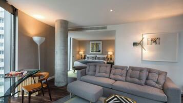 Una habitación de hotel moderna con un sofá gris, una mesa de vidrio y una silla. Hay una cama con cabecera y una mesita de noche con una lámpara. La habitación tiene un ventanal grande y vistas de la ciudad.